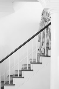 An older lady in B&W steps out of view at the top of some bannister stairs.