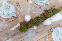 A bird's eye view of a table decorated for a wedding festivity. There's glassware, white candles, and place settings neatly arranged around a piece of greenery running down the middle of the table.