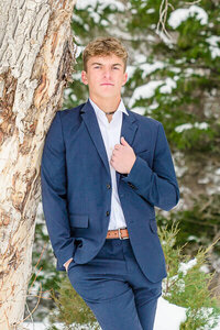 A high school senior boy wearing a dark blue suit with a brown leather belt leans agains a tree trunk at Tibble Fork Reservoir in the Winter. Captured by Salt Lake City Senior Photographer Melissa Woodruff Photography