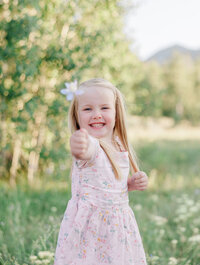 A happy toddler girl in a pink dress smiles big while showing off a wildflower 
