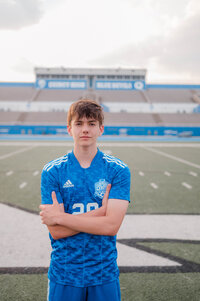 A teenage boy smiles at the camera with his arms crossed while standing on the soccer field.