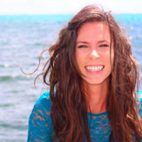 woman with curly brown hair and big smile standing with the ocean behind her