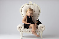 A charming studio portrait of a young child seated gracefully on an elegant, cream-colored throne chair. The child, dressed in a stylish black outfit, holds a small, smiling black bear plushie, radiating innocence and warmth. Captured by a skilled Dallas children's photographer.