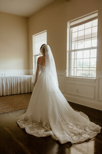 A bride in a long, flowing white gown and veil stands alone in a softly lit room with wooden floors and large windows, conveying a serene and contemplative mood.