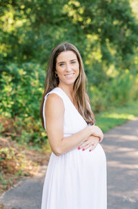 Expecting mom in white dress smiles at camera during Summer maternity session in Raleigh NC