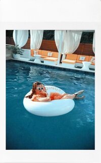 A woman in a coral dress lounges on a custom float in a luxurious pool surrounded by cabanas, creating a relaxed and playful vibe.