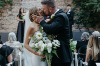 Bride and groom sharing a kiss at their wedding ceremony, holding a bouquet of white flowers, in an outdoor setting at Margaret Place in New Orleans.