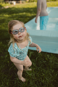 toddler girl looks up to the camera while wearing goggles standing next to a large inflatable pool