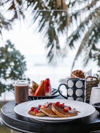 Beach view with a plate of pancakes topped with fruits. Side of coffee and more fruits