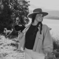 Photographer smiling while holding camera outdoors with mountains and lake in the background.