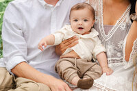 A baby, wearing a tan outfit, sits on his mom and dad's laps and smiles at the camera.