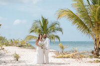 a couple stands by a palm tree near the ocean on their wedding day