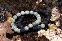 A Moss Agate Bracelet Lays on Top of a Lava Rock Bracelet with the Ocean in the background.