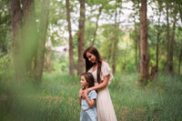 A mother has her arms on her daughter in front of her and she looks down at her and smiles. They are in a beautiful green park in Kamloops, BC.