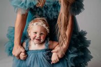 A young child, standing in front of her mother as her mom holds onto her hands. You can't see much of her mom, but the are wearing matching blue tulle gowns and looks so happy to be together. The toddler is also wearing a white floral headband.