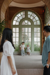 Two children in matching outfits framed by a tall arched window, as their parents look on lovingly.