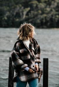 A person standing by the water's edge, gazing into the distance, wrapped in the comfort of a chic, oversized plaid coat that suggests a crisp, serene day.