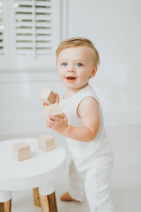 Baby boy in white romper holds wooden blocks and plays during photography session in Raleigh NC