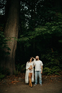 Expecting couple stands beneath tall evergreens during a forest maternity photoshoot; the woman wears a sheer robe and boots, cradling her belly, while the man gently touches her hair.