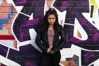 A feminine presenting teenager wearing a black leather jacket and long hair pulled up in a purple scrunchie leans against a wall, looking at the camera with a serious expression. The wall behind them is painted with a purple, white, and orange graffiti mural.