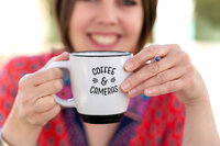 Kate Daniel, a Calgary brand photographer, holds a white enamel coffee mug with ‘Coffee & Cameras’ printed on it. The focus is on the mug and her hands, adorned with a simple ring, while her smiling face is softly blurred in the background. She wears a vibrant, patterned top, adding a warm and creative touch to the image.