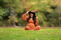 Pregnant mother sitting on the ground for a fall maternity photo session wearing an orange gown and hodling on to her large brimmed brown hat