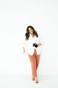 Client of Bostron Design Studio, Adriana holds her camera in one hand and is smiling with her head slightly tilted. She is in a white studio space.