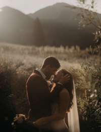 A bride and groom are about to kiss while standing in a meadow at the foot of a mountain. The groom has his hand on the side of the bride's head
