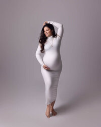 maternity mom wearing white dress on a white backdrop. Hand on belly and hand on head pose.
