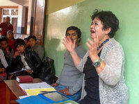 Nita Tin speaking at Timothy Initiative gathering in Kathmandu, Nepal, smiling brightly with hands raised as she addresses church leaders and pastors. Nepalese church leaders sit beside her listening intently. She wears a black dress with a white sweater, a bead necklace, and a silver watch.