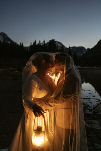A bride and groom share an intimate moment beneath a sheer veil, illuminated by the warm glow of a lantern beside a mountain lake at dusk, captured by Sydney Breann Photography.