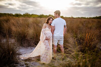 Woman wearing flowing lace maternity dress. She is holding her pregnancy belly with snuggling with husband. Captured during golden hour at Cypress Point Park in Tampa, FL