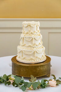 A three-tiered white wedding cake with ornate icing decorations sits on a gold cake stand, placed on a white table with green eucalyptus leaves and a pale pink rose.