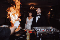 A sample image from Philadelphia wedding photographer Daring Romantics. A couple watches in excitement as the fire of cooking food roars in front of them.