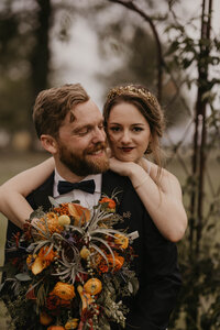 Bride and groom pose for photo, as groom sits and smiles off to the side while bride bends down and throws her bouquet over her new husband and smiles sweetly at the camera