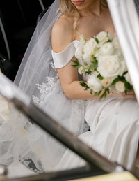 Bride sitting in a car, holding a bouquet of white roses. She wears an off-shoulder dress and lace veil, exuding elegance and anticipation.
