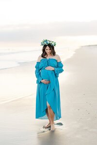 pregnant mom holding baby bump at Emerald Isle Beach in blue dress with flower crown at sunset