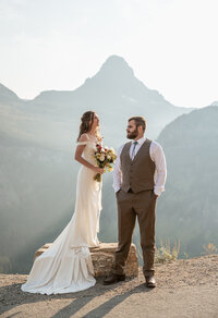 A bride and groom stand together overlooking mountain peaks in Glacier National Park, illuminated by soft sunrise light, captured by Sydney Breann Photography during their Montana elopement.