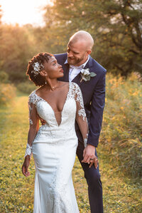 Bride with tight curls and unique hair piece and lace low cut wedding gown and bold red lip stick looks up at groom in blue suit jacket with white boutonniere while standing in wild flowers during golden hour in Wisconsin