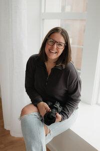 A photographer smiling as they sit by a window holding a camera 