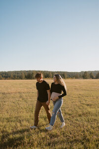 A husband holding hands with his pregnant wife, walking in a grass field outside.