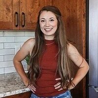 A woman with long brown hair, wearing a sleeveless red top and blue jeans, stands in a kitchen smiling at the camera, as the Virtual Assistant of a Shreveport website design company.