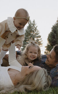 A wife holds up their baby smiling. Her husband is next to her and their toddler is on top of him. They are all lying on the ground in a park.