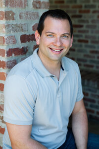 Headshot photo of a handsome business man with a light gray collared shirt smiling by a brick wall in San Juan Capistrano, CA.