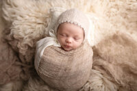 Baby girl wrapped in a neutral blanket wearing a cream colored bonnet.