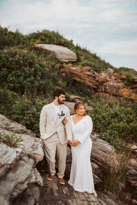 A couple holds hands on the rocks with green foliage and roses behind them.