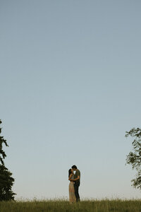 Engaged couple embracing on a grassy hilltop under a clear blue sky, captured in a natural, documentary style by a Michigan wedding photographer.