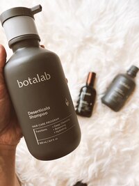 Hand holding botalab Deserticola Shampoo for deep cleansing in front of other riman hair care products that are blurred in the background