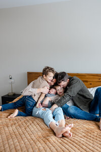 a family of 5 snuggles on a bed with a brown comforter as they all squeeze eachother in a hug, smiling and laughing