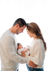 Parents with facing each other, with temples touching, close their eyes as they carry a newborn baby girl in between them.